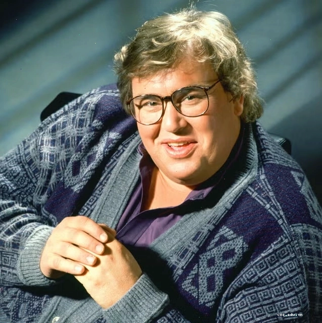 John Candy ngồi trên ghế, mỉm cười và mặc áo len.